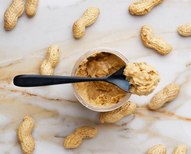 Overhead shot of a jar of peanut butter surrounded by peanuts in the shell. A black spoon with peanut butter in it is laying across the jar