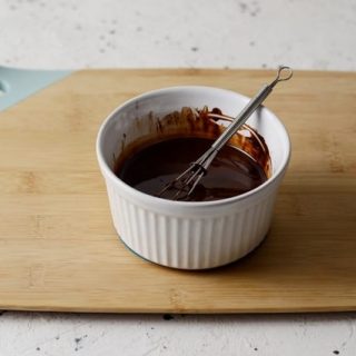 White ramekin of chocolate sauce with a small whisk in it, sitting on a bamboo cutting board with marble backdrop