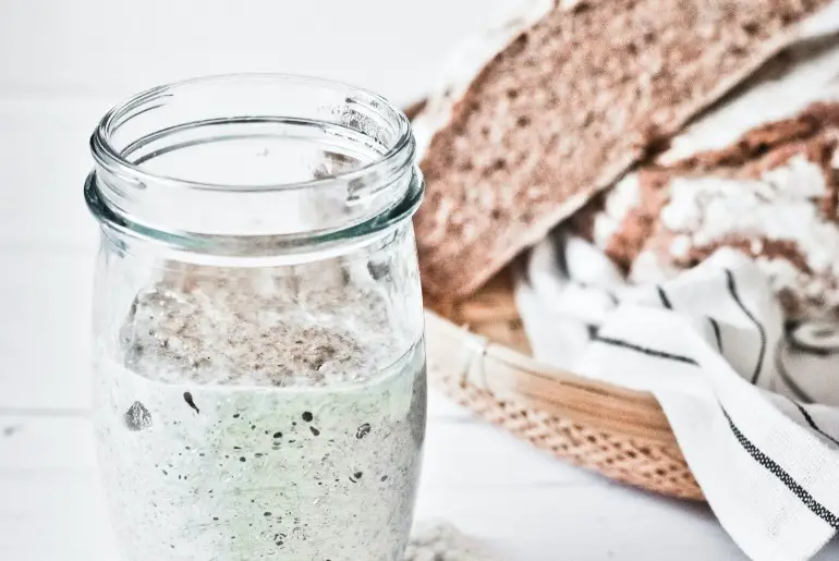 sourdough starter in the foreground with a basket of sourdough bread in the background