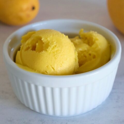 vibrant yellow-orange ice cream in a white ramekin with 2 mangos in the background