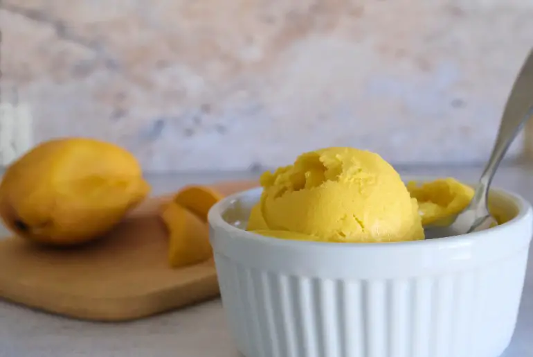 vibrant yellow ice cream in a white ramekin with a half cut up mango in the background