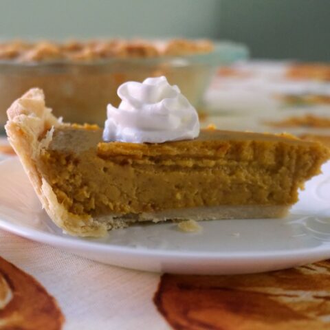 slice of pumpkin pie on a white plate with a pie in a glass dish behind it