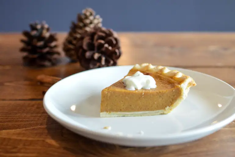 a slice of pumpkin pie on a white plate with pinecones in the background