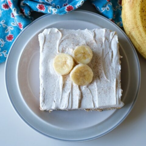 square banana cake on a pale green plate. The cake is golden brown with white, fluffy frosting and 3 banana slices on top. There are several bananas and a blue floral towel at the top of the image.