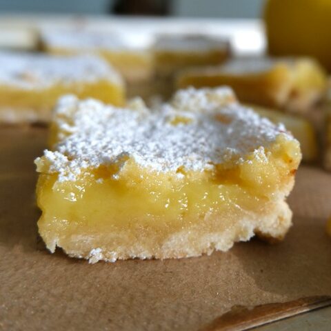 A single lemon bar placed on brown parchment paper, with additional lemon bars visible in the background. The lemon bar is square-shaped with a golden brown crust and a vibrant yellow lemon filling that is visible in the center.