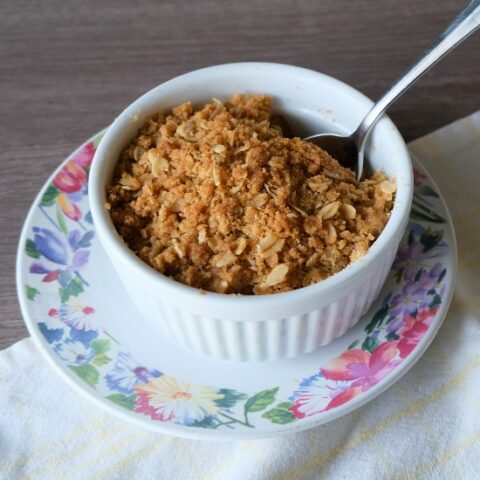 brown oatmeal crisp in a white ramekin with a spoon in it. The ramekin is sitting on a floral plate and sitting on a pale yellow cloth