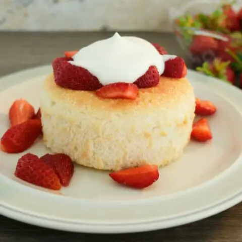 small, round, pale yellow cake on a plain plate topped with sliced strawberries and a dollop of whipped cream. A small bowl of strawberries is in the background.