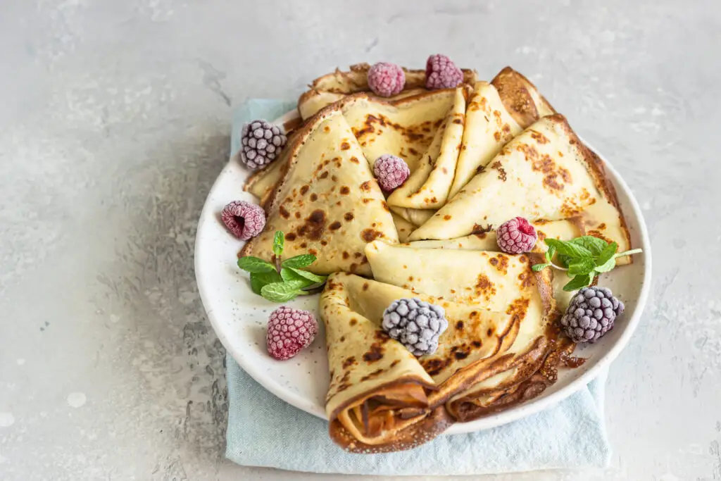 Several crepes fill a small white plate and are topped with red and purple berries and green mint leaves.