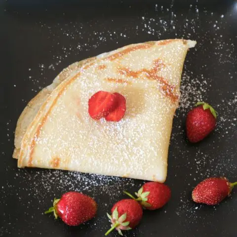 lightly golden crepe folded into a triangle on black plate. The crepe is dusted with powdered sugar and surrounded by red strawberries
