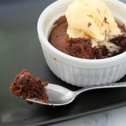 A brownie in a white ramekin sits on a black plate. The brownie is topped with ice cream. A silver spoon with a bite of brownie rests on the plate in front of the ramekin.