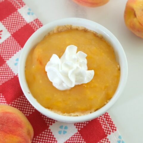 image features a mini peach pie in a white ramekin on a red and white checkered cloth. There are several peaches around the pie and the pie is topped with a small dollop of whipped cream.